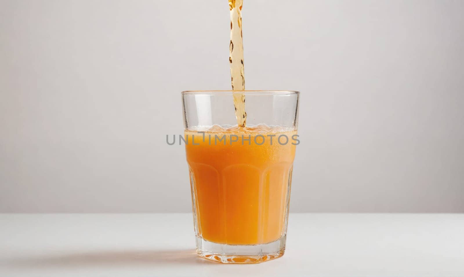 Orange juice is being poured into a clear glass. The glass is sitting on a white table. The juice is a bright orange color and is creating a small amount of foam as it pours.