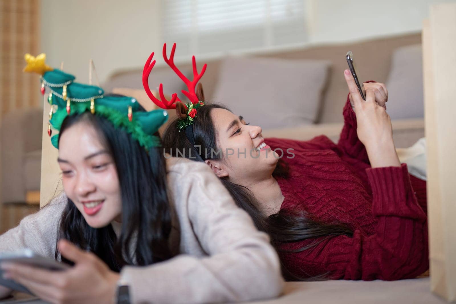 Lesbian friends wearing festive headpieces engage in joyful smartphone connection amidst Christmas decorations, celebrating inclusivity and holiday cheer.