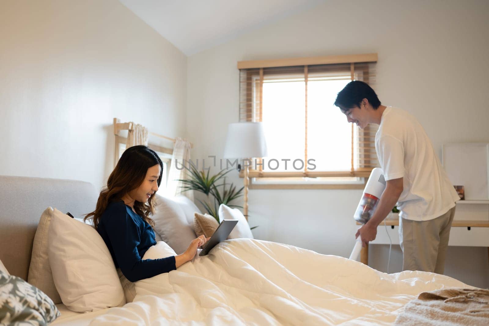 Millennial woman using digital tablet on bed while her husband vacuuming floor in bedroom.