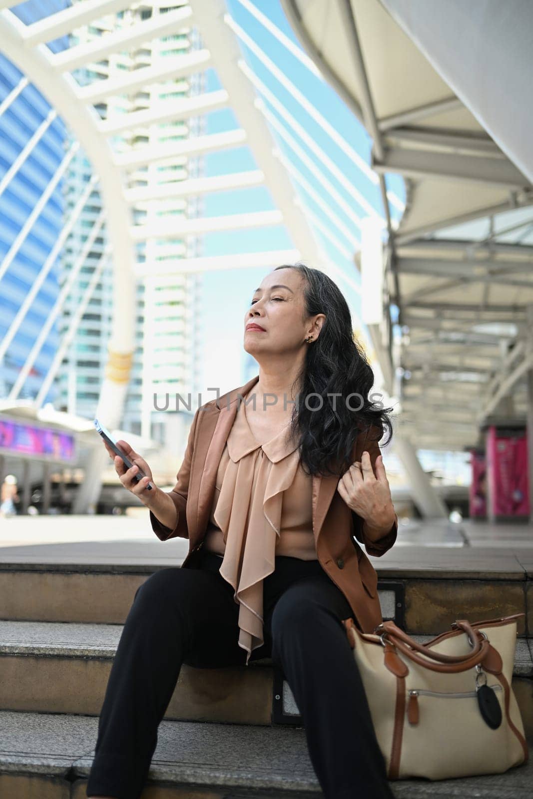 Portrait of elegant senior businesswoman relaxing on the steps in modern cityscape.