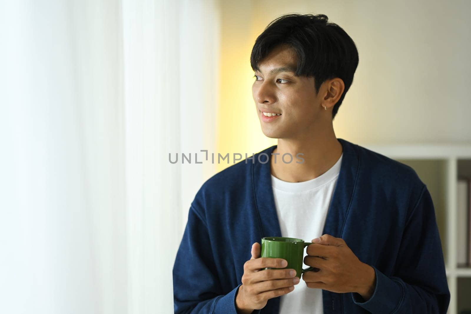 Relaxed young adult man holding a coffee cup gazing outside with a smile.