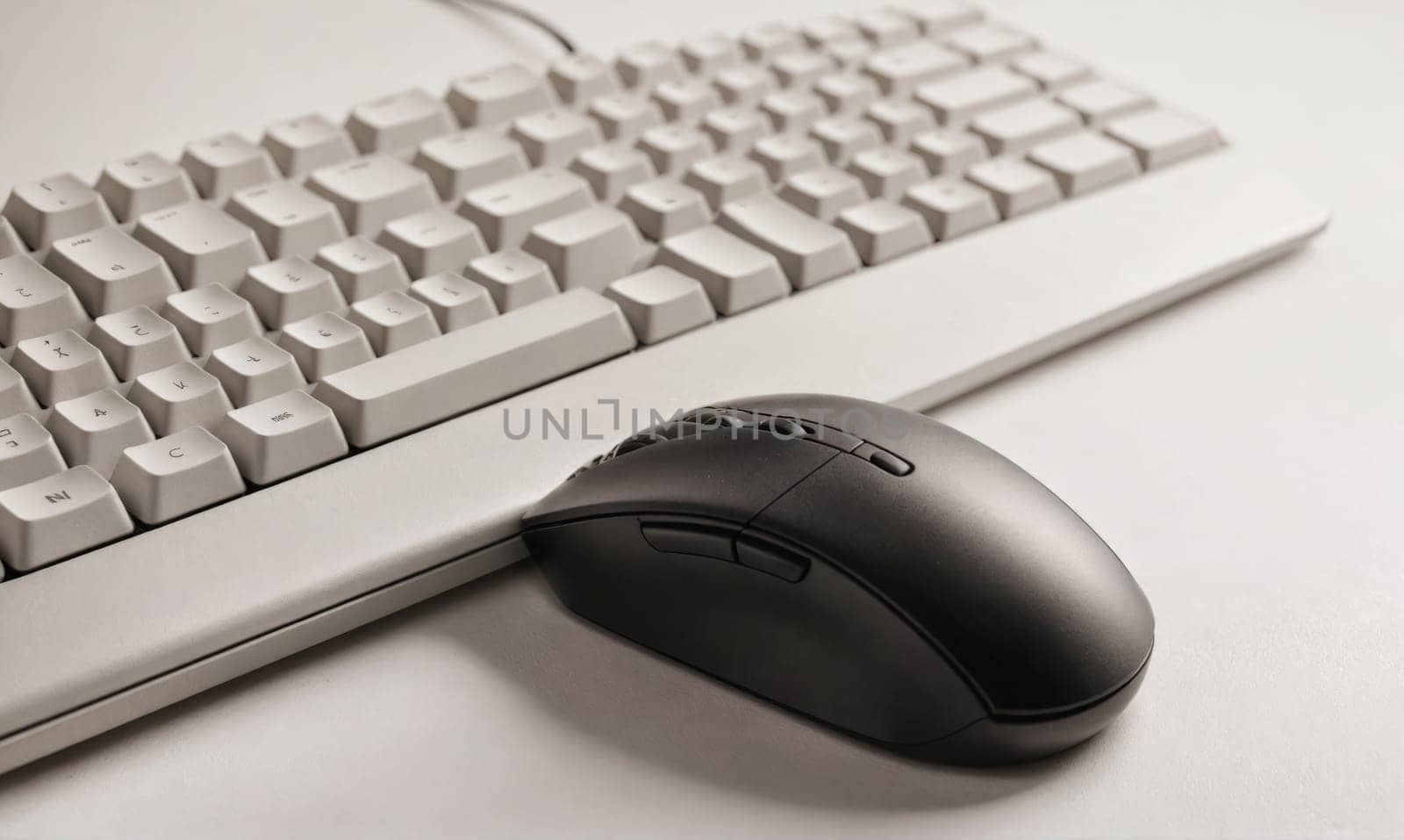 A white keyboard lies on a white desk, with a black mouse sitting on the desk below it.