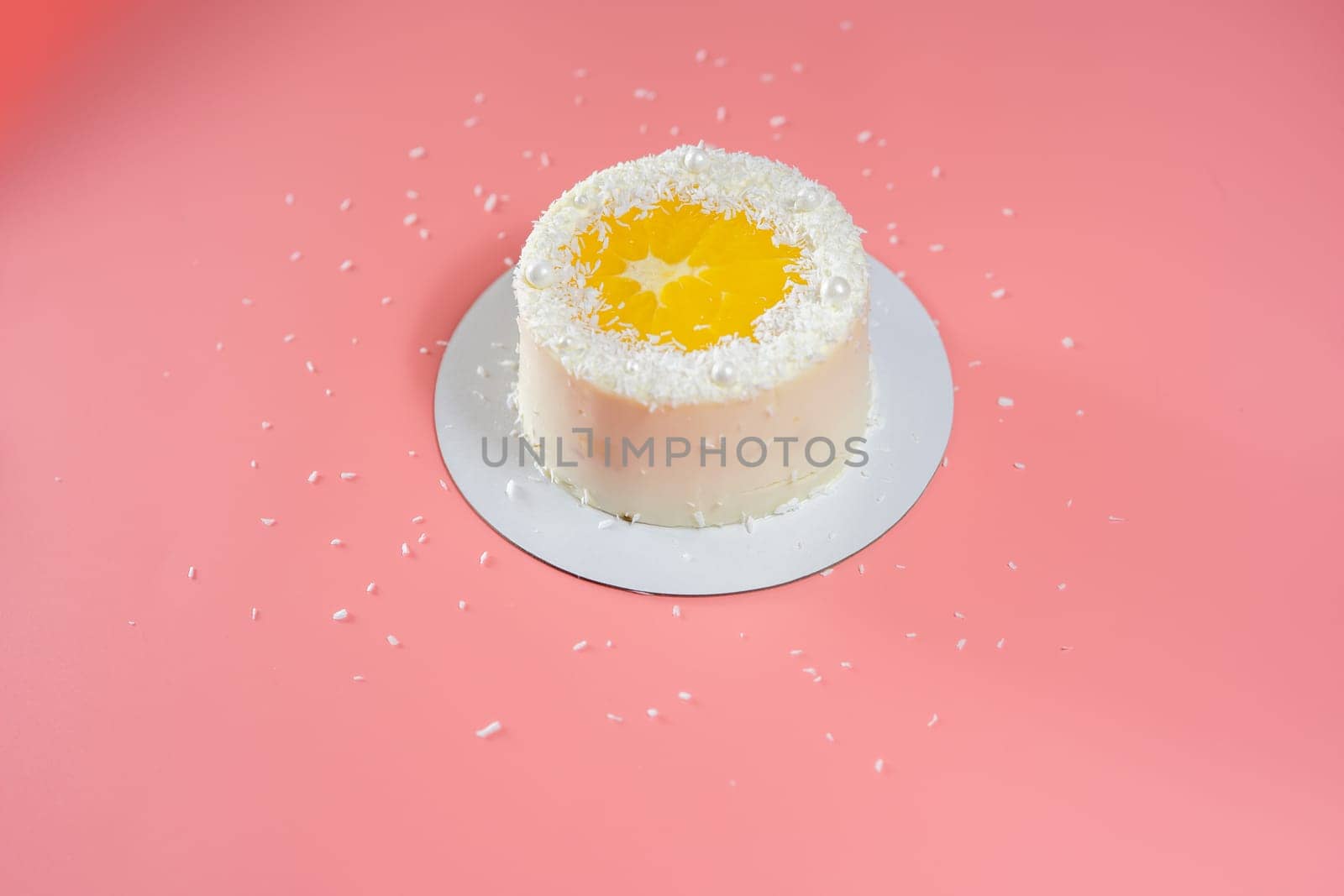 Delicious white cake decorated with orange slice and coconut flakes standing on pink background with scattered coconut flakes