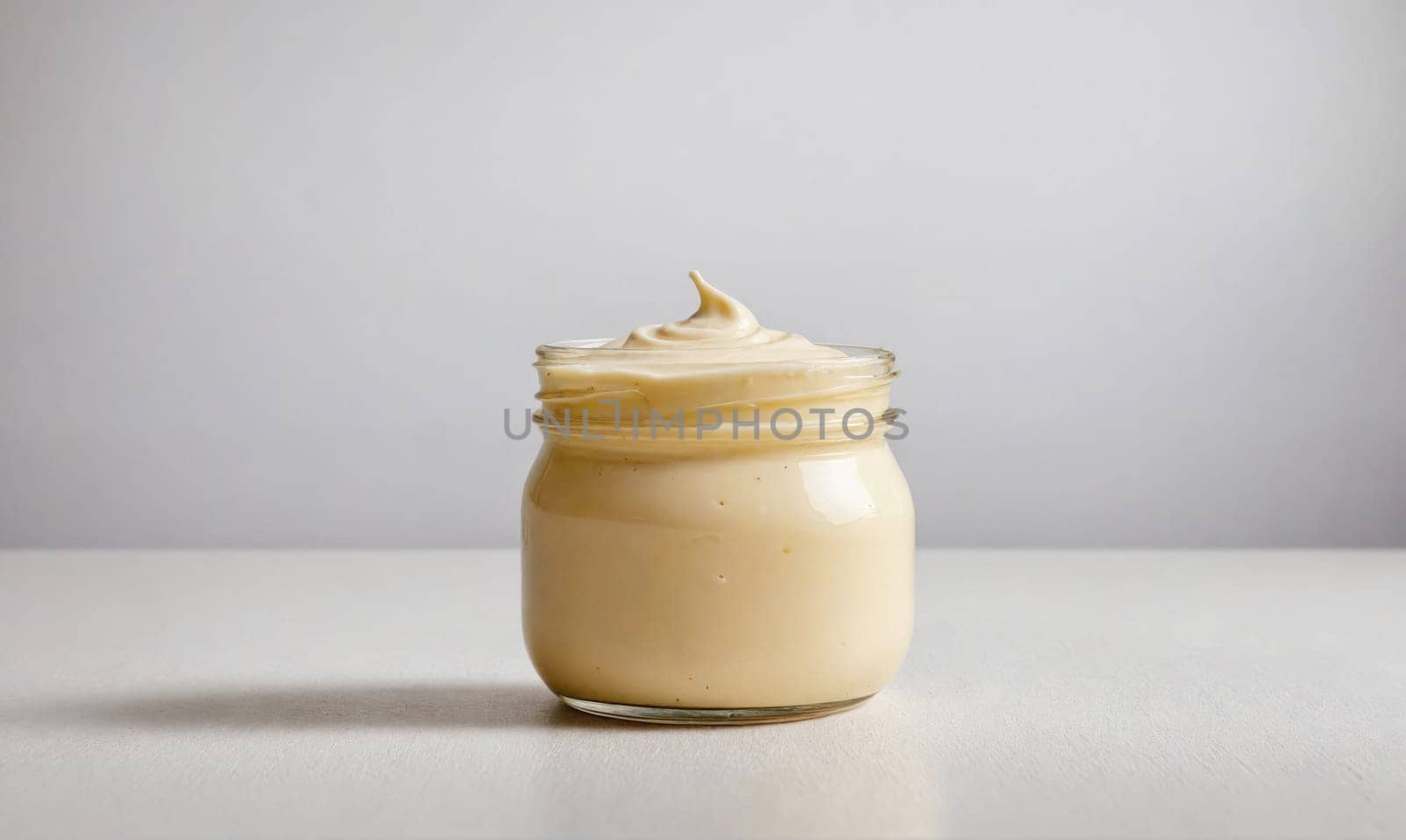 A glass jar filled with thick, creamy, light-colored sauce is positioned on a white surface. The sauce has a swirled top.