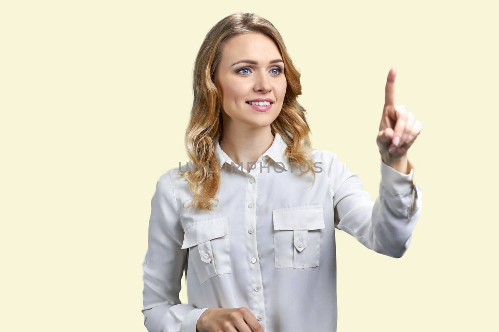 Smiling blond woman is touching transparent virtual screen. Pastel yellow background.