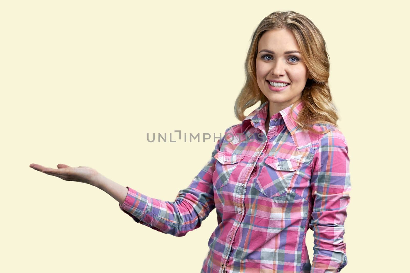 Smiling young blond woman is showing copy space with one hand. Isolated on pastel yellow background.