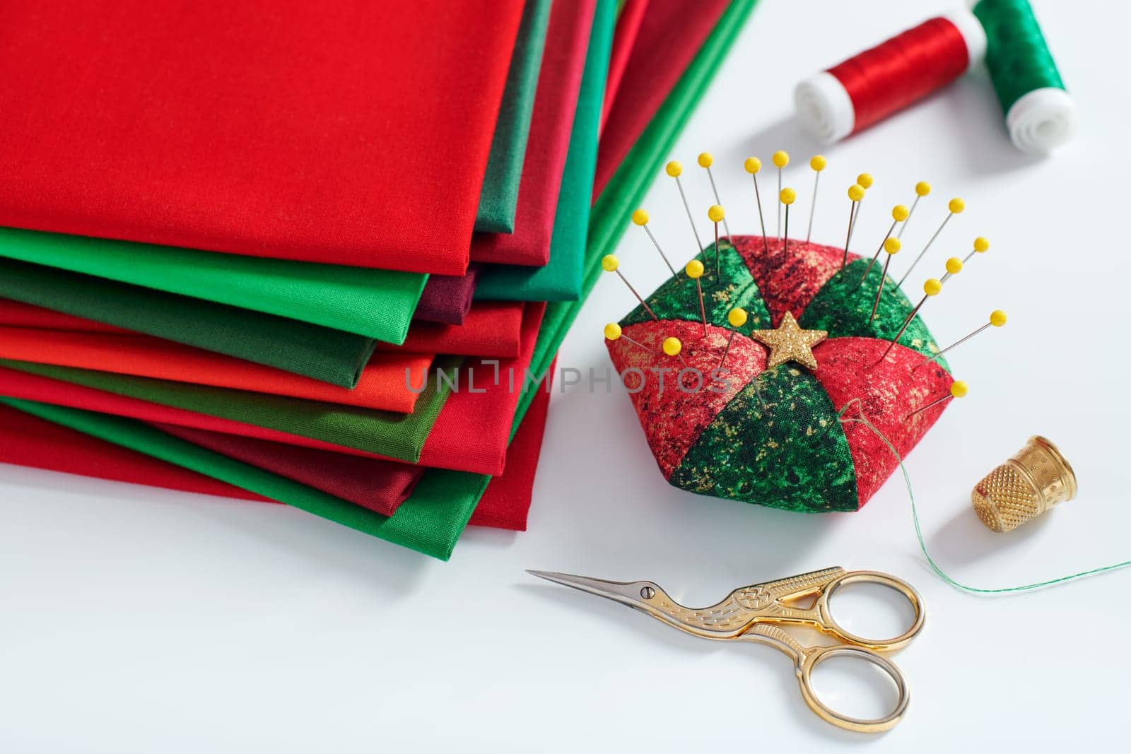 Pile of red and green fabrics, pincushion, scissors, thimble and spools of thread on white background