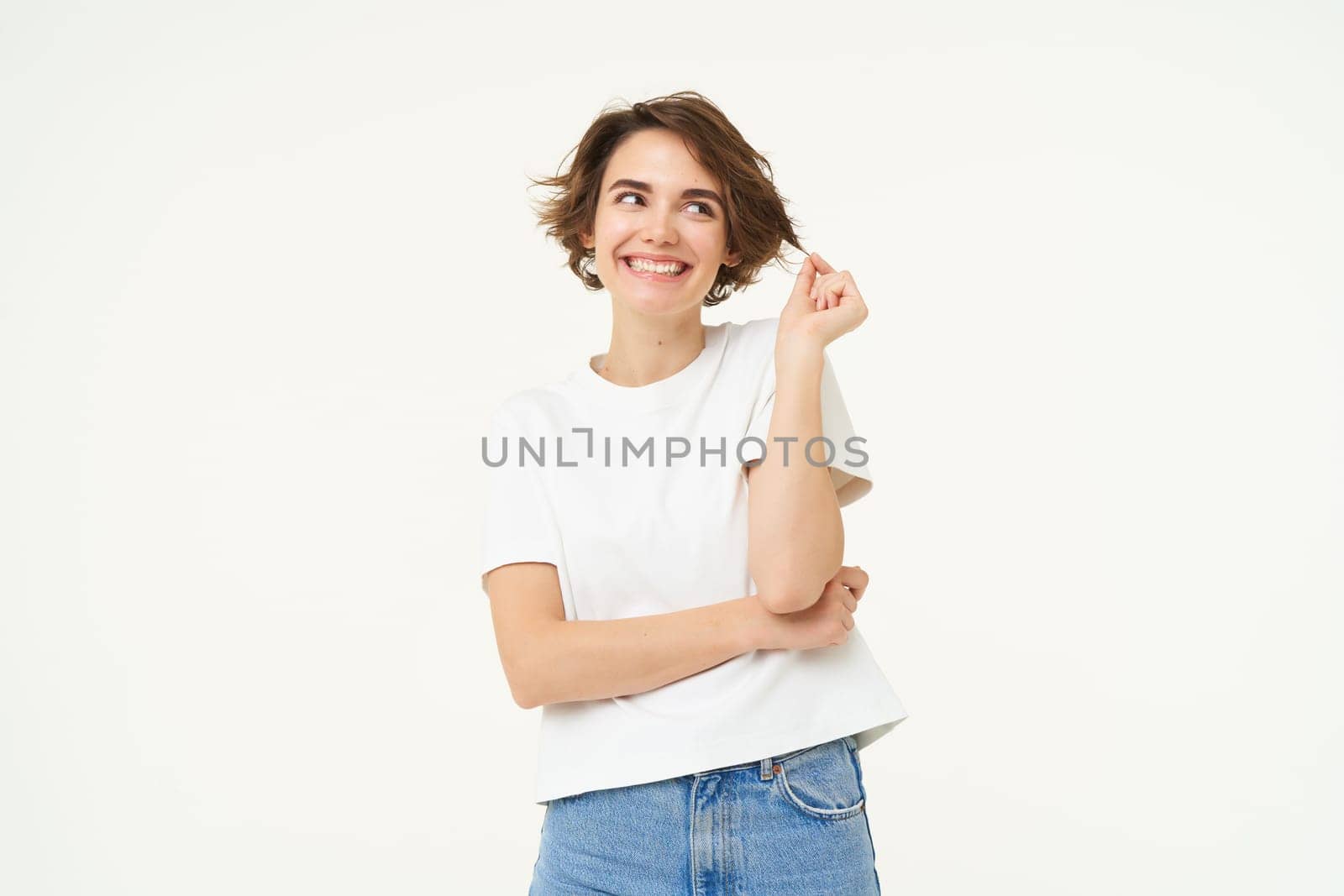 Portrait of confident woman with short hair, playing with strand, smiling and looking aside with thoughtful look, standing over white background. Copy space