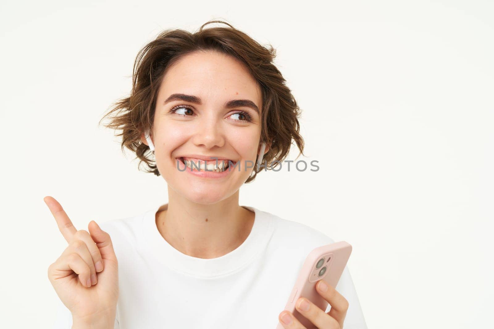 Portrait of brunette woman listening music in headphones, pointing left at banner, holding mobile phone, using streaming app, isolated over white background.