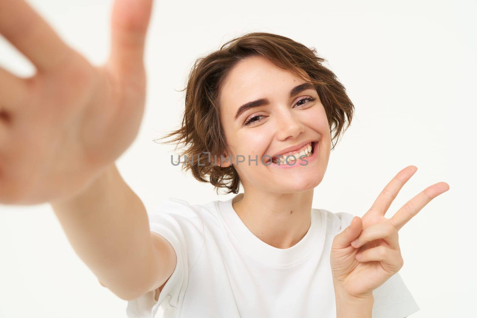 Lifestyle and people. Young woman smiling and taking selfie, posing for photo, holding camera with one hand, standing over white background.