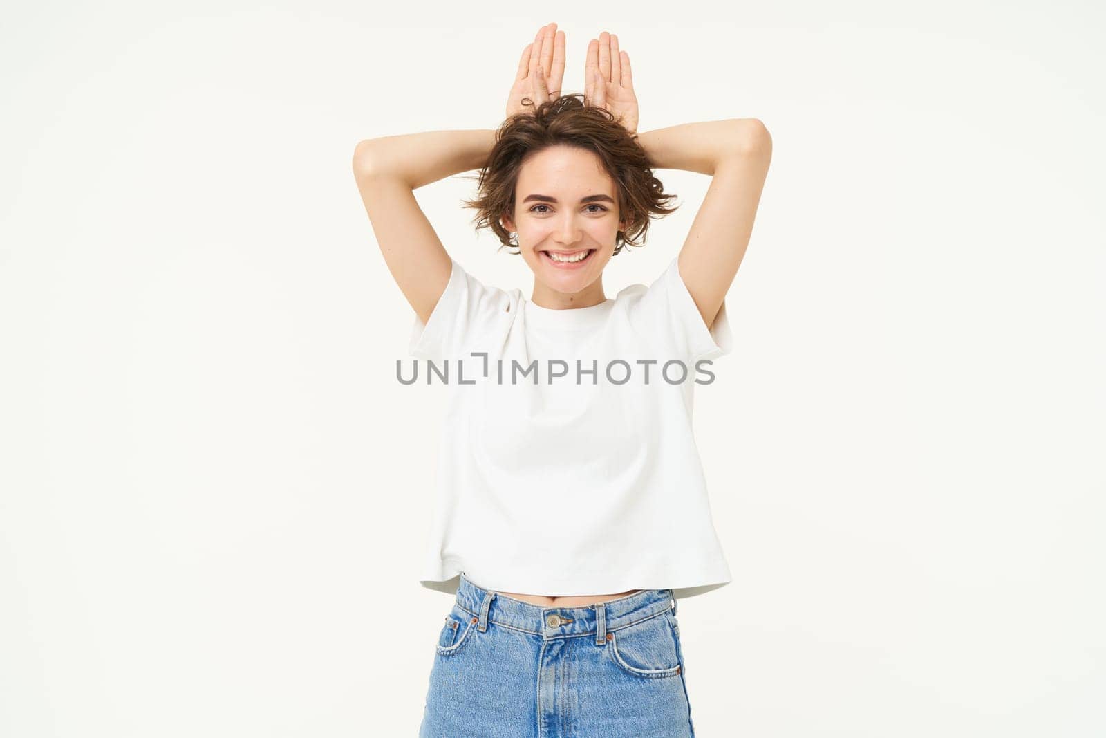 Portrait of funny and cute young woman with bunny ears gesture, holding palms on top of her head, smiling and looking happy at camera, standing over white background.