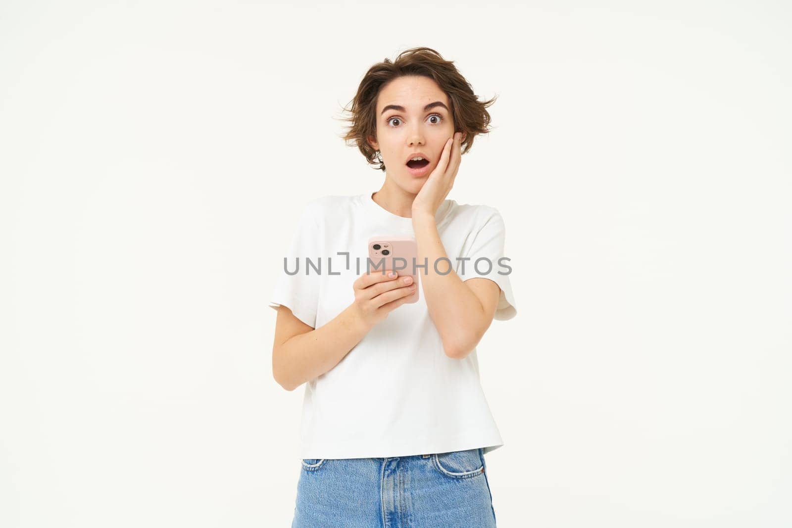 Portrait of shocked young woman with smartphone, drops jaw and says wow, reads big news on mobile phone app, isolated over white background.