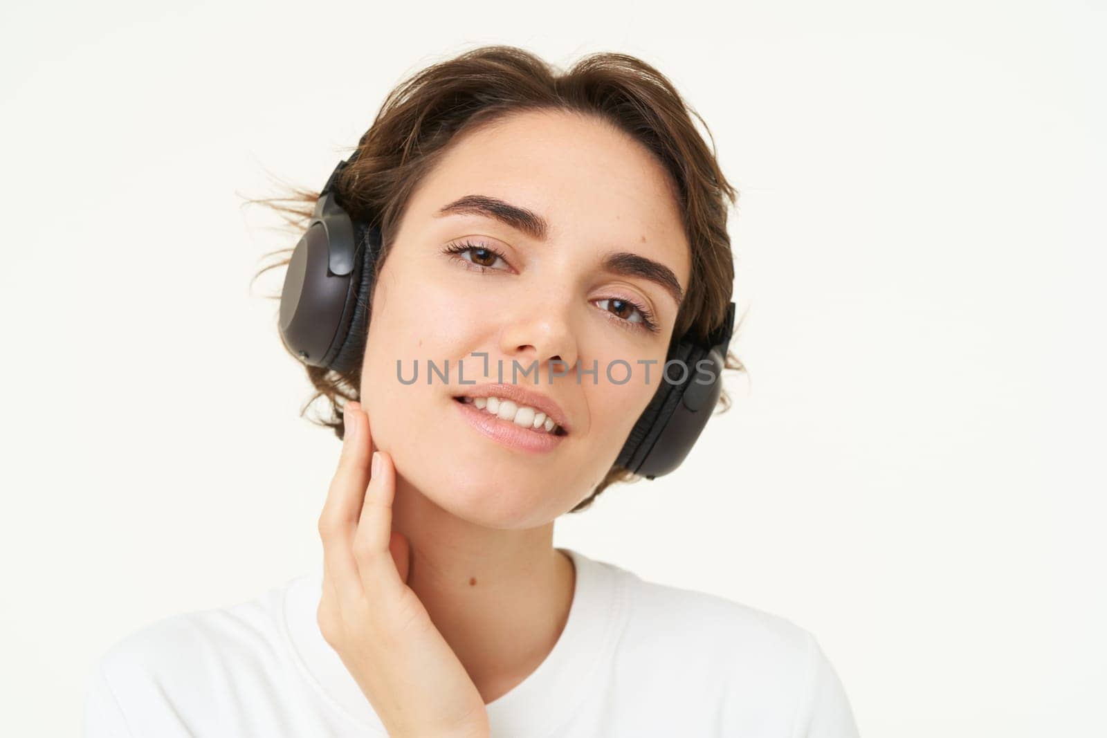 Portrait of young candid woman in wireless headphones, smiling, listening music in earphones, standing over white background.