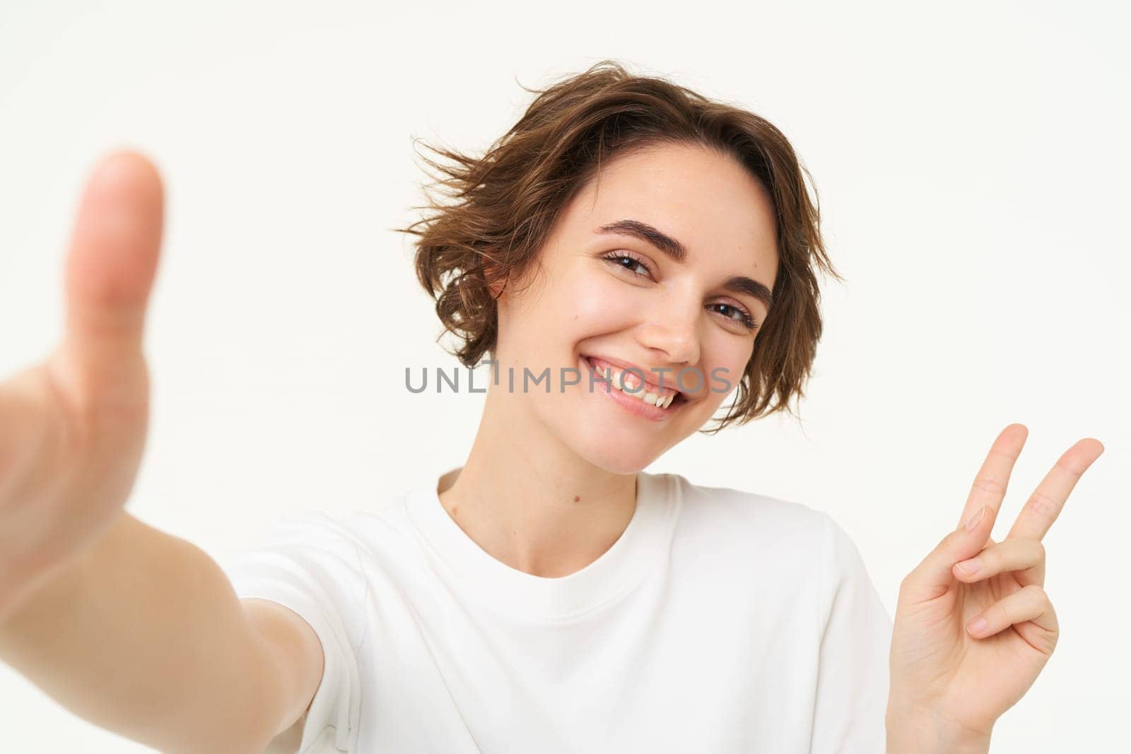 Close up of smiling brunette woman makes selfie, taking photo for her social media, holds mobile phone with extended hand, standing over white background.