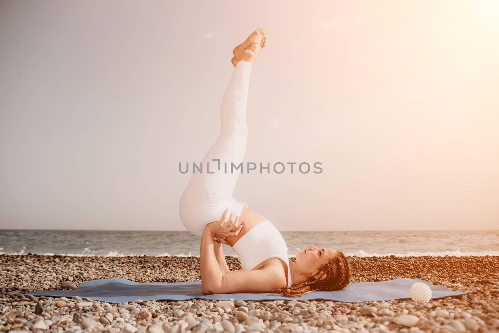 Woman yoga sea. Well looking middle aged woman with braids dreadlocks in white leggings and tops doing stretching pilates on yoga mat near sea. Female fitness yoga routine concept. Healthy lifestyle