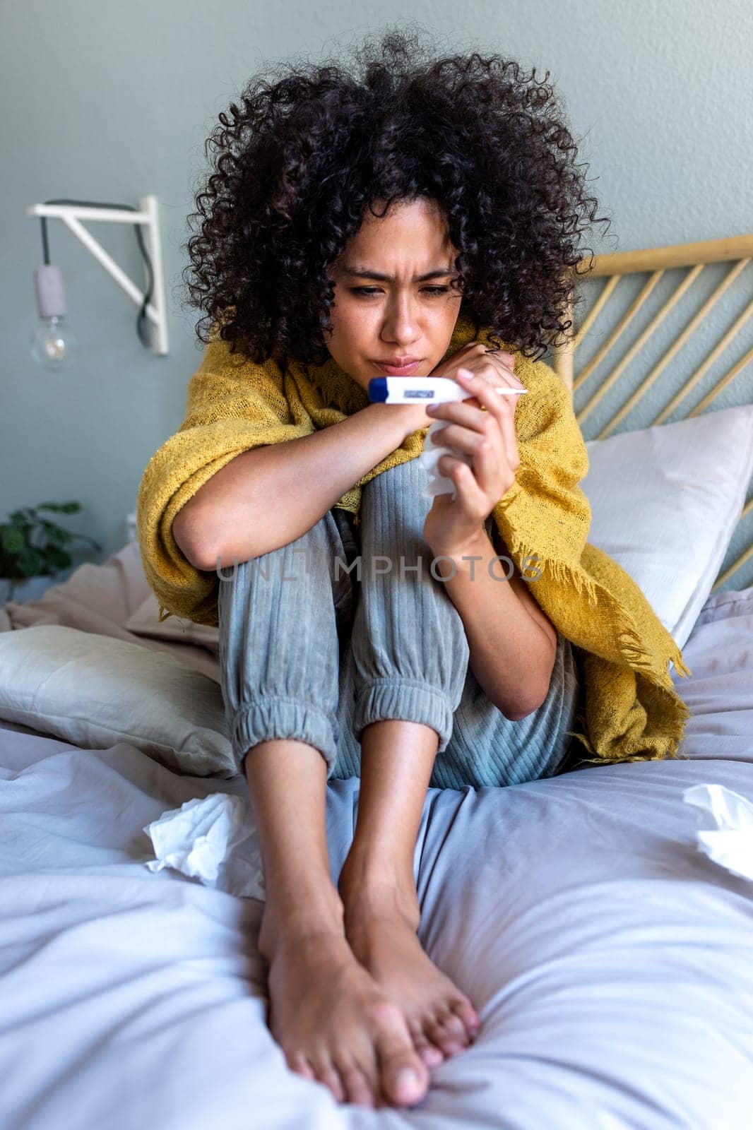 Multiracial woman with curly hair having the flu checking temperature with digital thermometer. Vertical image. by Hoverstock