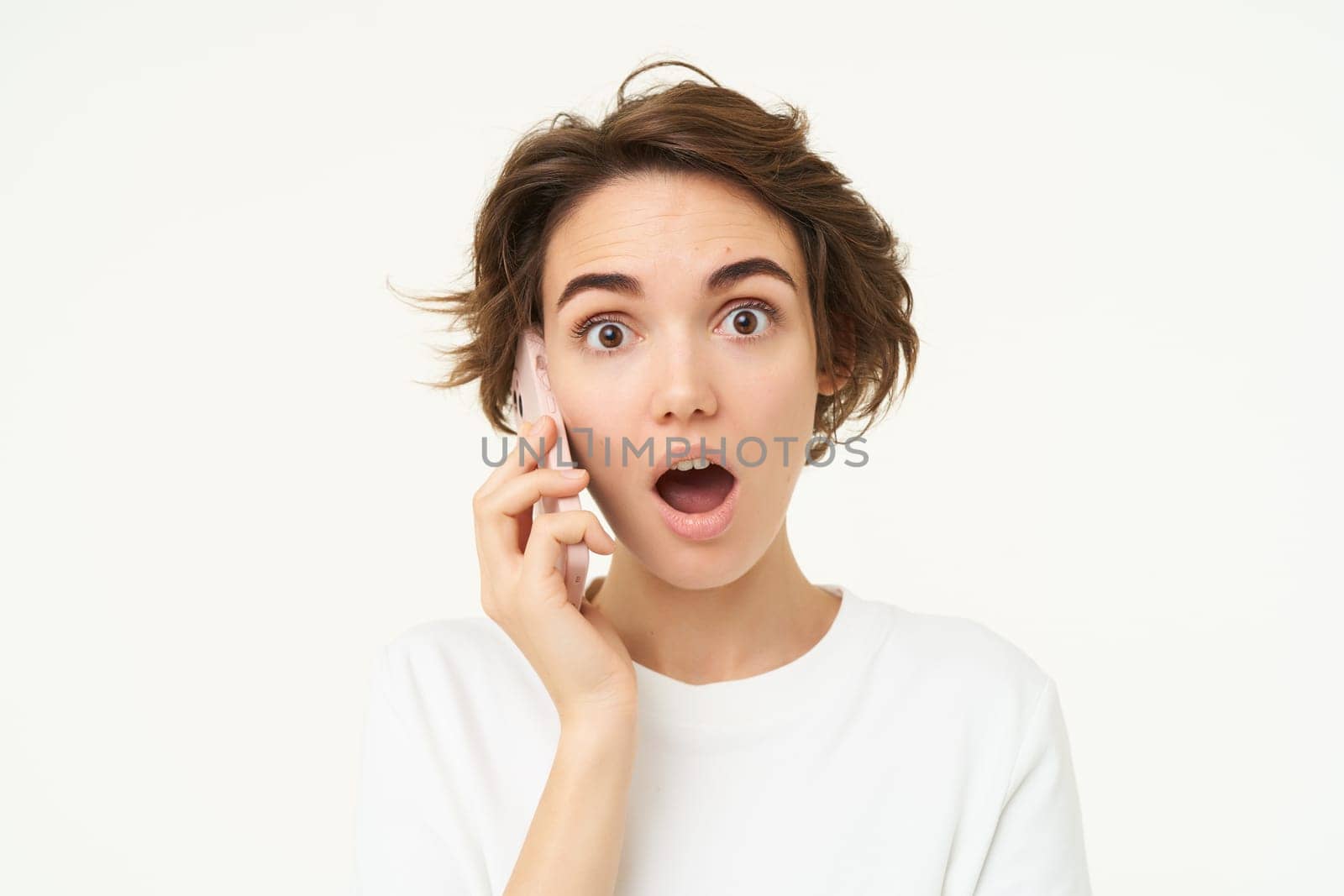 Portrait of woman with surprised face, answers phone call and looks excited, amazed by big news, stands over white background.