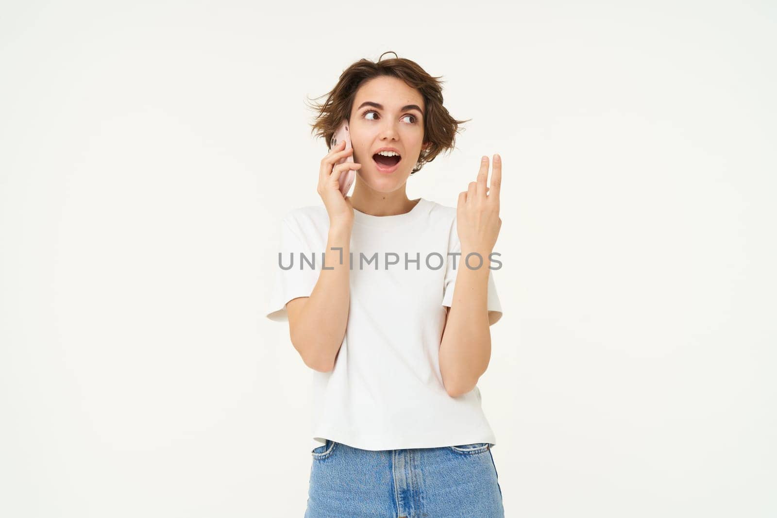 Portrait of chatty young woman, talking on mobile phone and gesturing, discussing something over the telephone conversation, standing over white background.