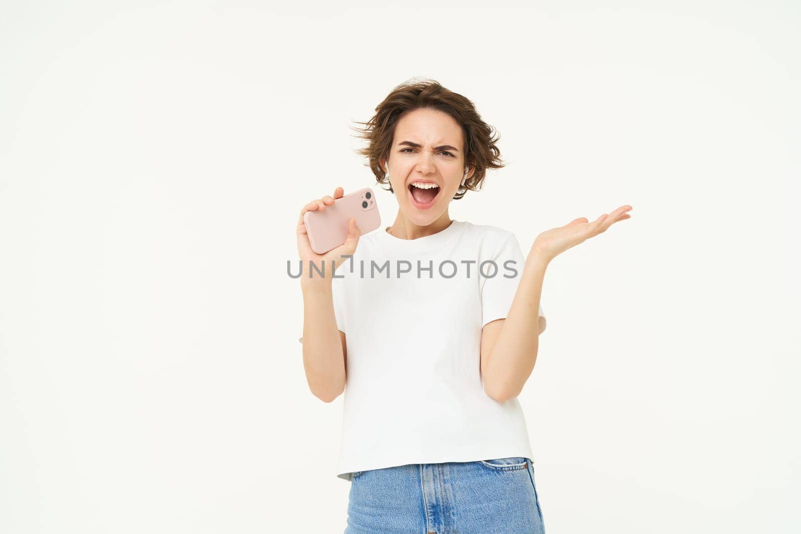 Portrait of carefree girl singing in wireless headphones into smartphone microphone, playing music game on mobile app, standing over white background.