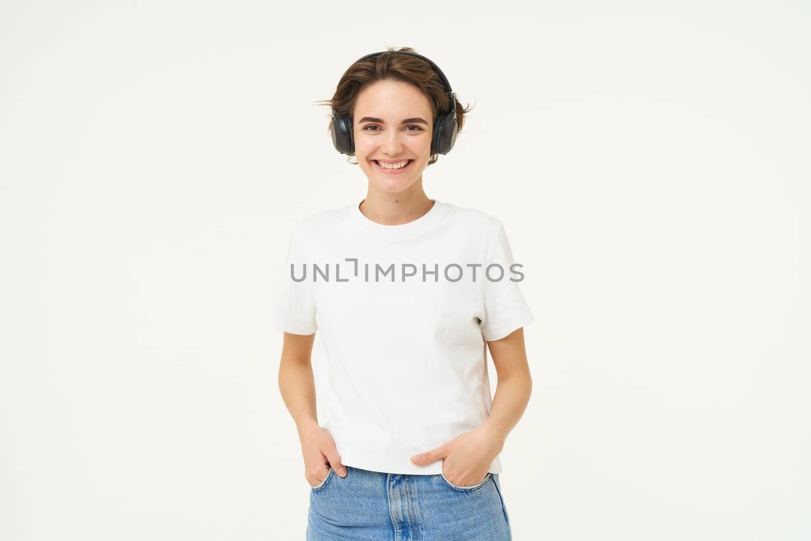 Image of modern girl in wireless headphones, listens to music in earphones, smiles and looks happy, poses in casual clothes over white background.