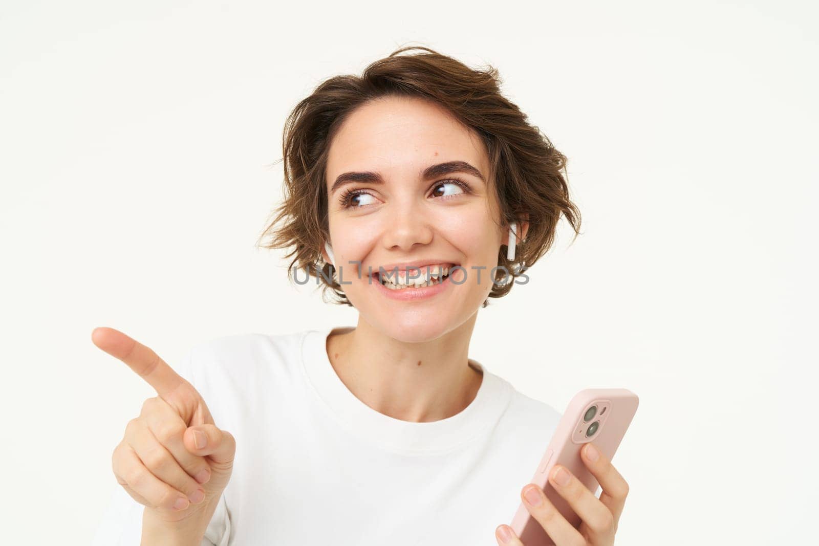 Woman in wireless headphones, holding smartphone, pointing left and smiling, listening to podcast or music, white background by Benzoix