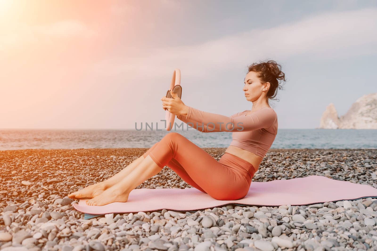 Woman sea pilates. Sporty happy middle aged woman practicing fitness on beach near sea, smiling active female training with ring on yoga mat outside, enjoying healthy lifestyle, harmony and meditation by panophotograph