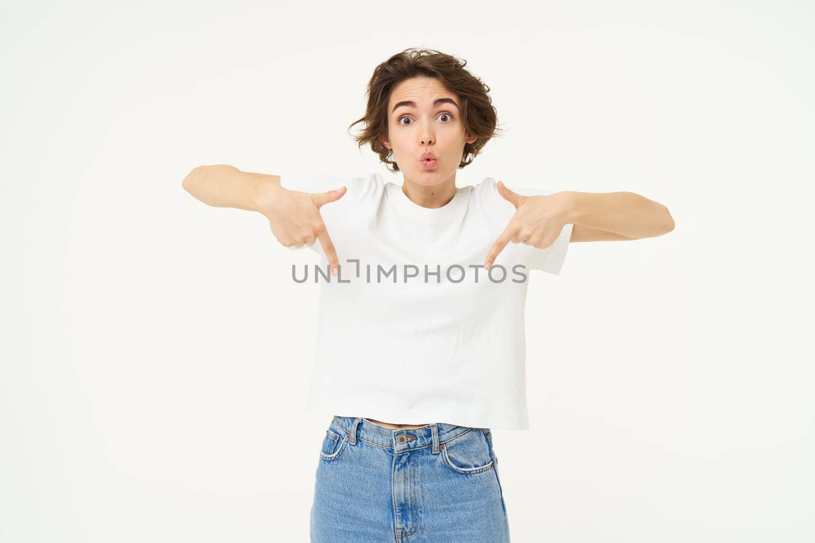 Portrait of amazed girl, pointing fingers down, showing something and looking surprised, standing over white studio background by Benzoix