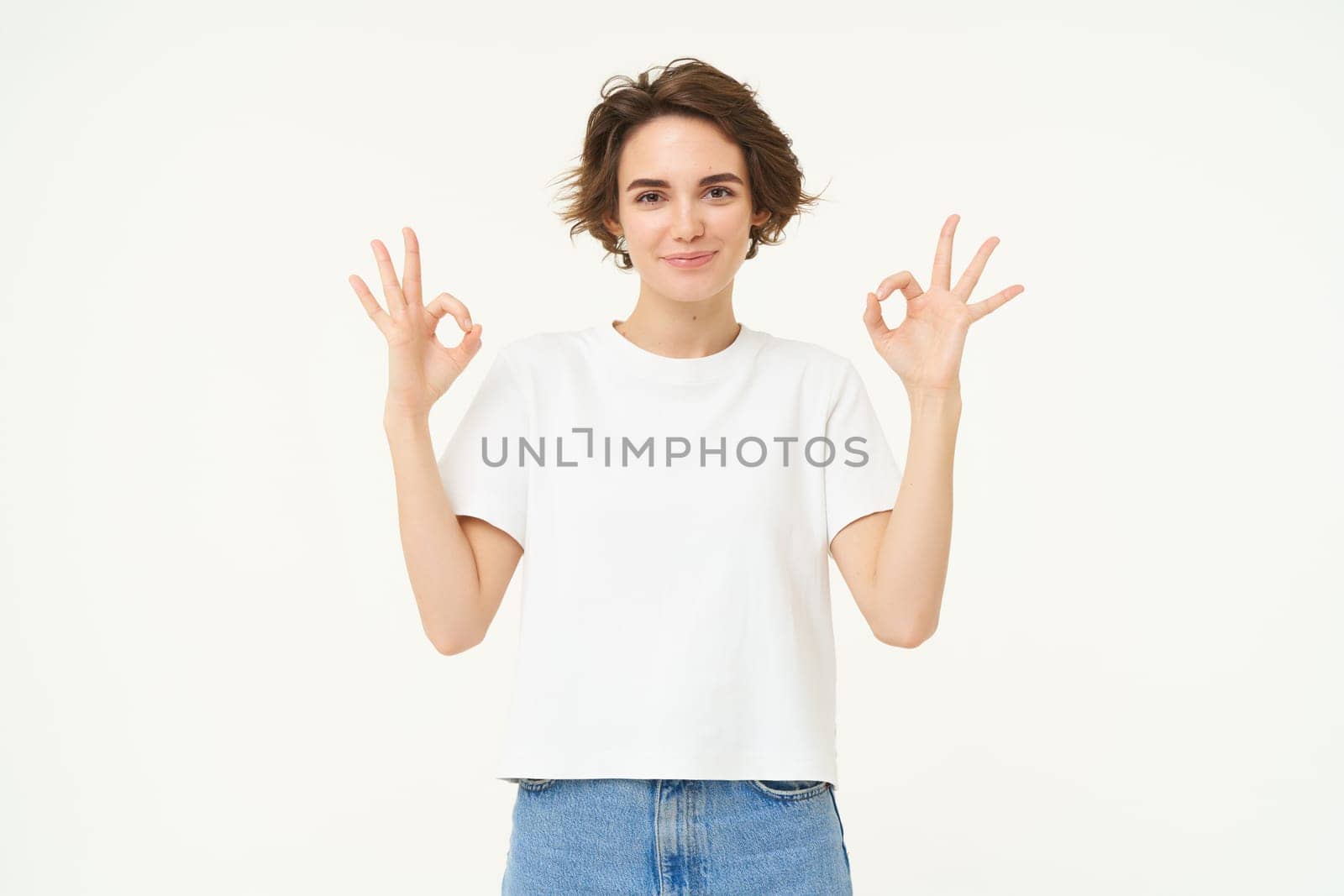 Portrait of young brunette woman, look confident, shows okay, ok sign, approves something, recommending, give positive feedback, stands over white background.