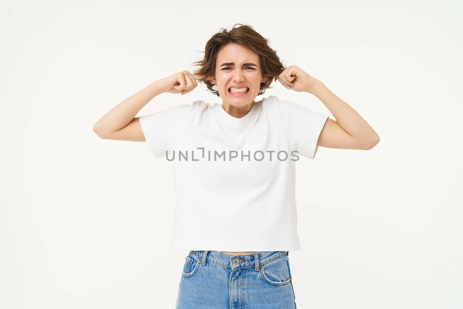 Portrait of upset woman shuts her ears, feeling discomfort from loud noise, standing over white background. Copy space