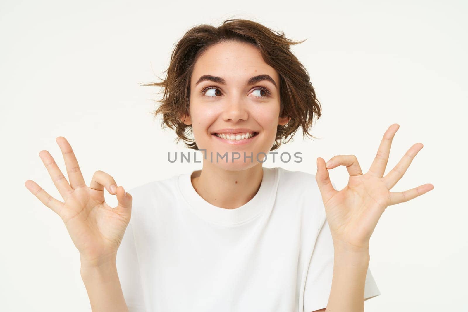 Image of excited brunette woman, smiles, shows okay, ok gesture, looks amused and happy, stands over white studio background.