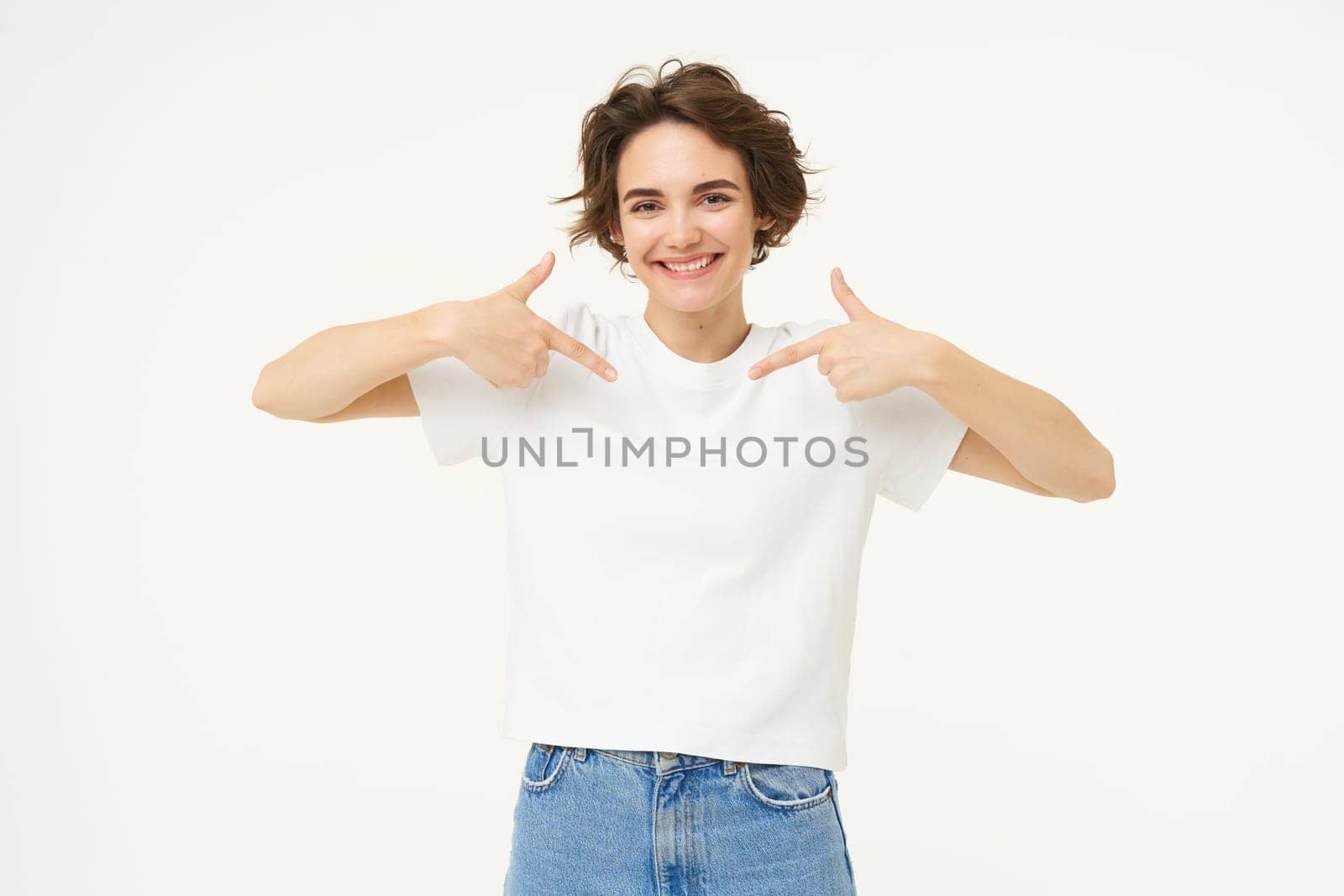 Image of confident, good-looking young woman, smiling and pointing at herself, standing over white studio background. Copy space