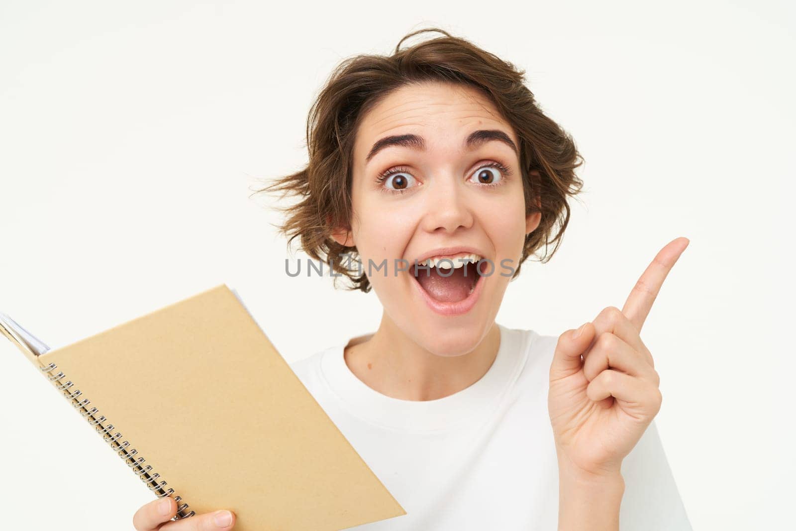 Enthusiastic brunette woman with diary, holding planner book, pointing at upper right corner and smiling with excitement, standing over white background.