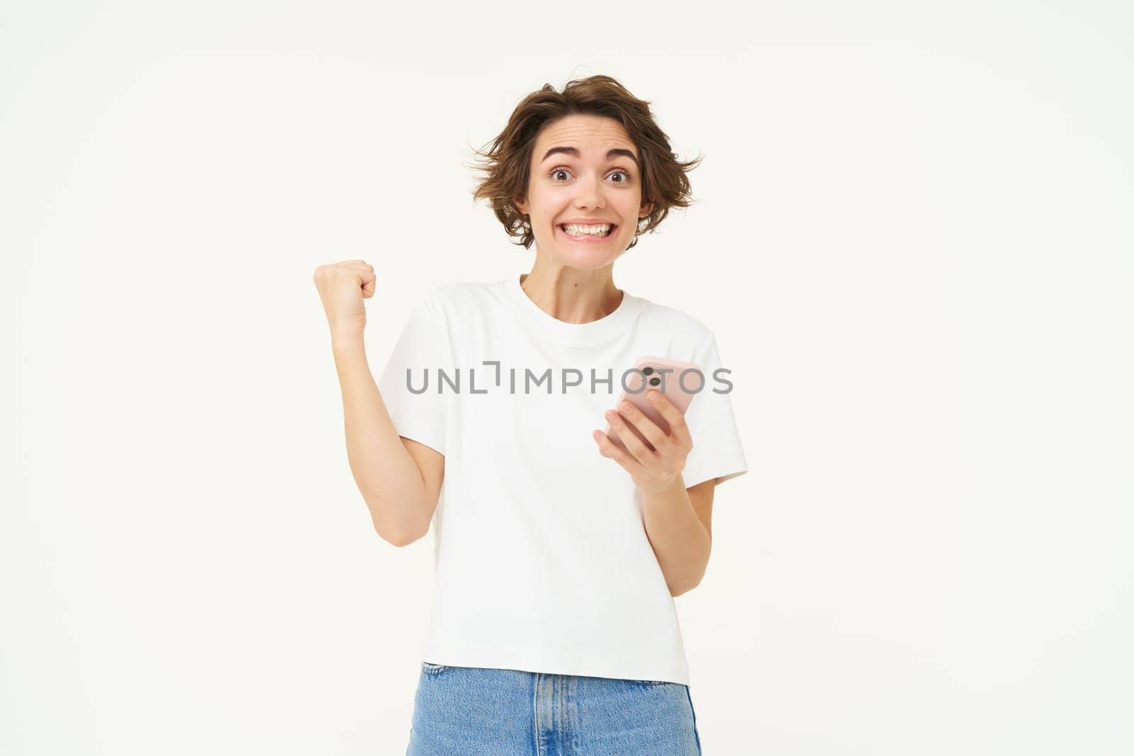 Portrait of excited brunette woman, holding smartphone and celebrating big win, achieves goal in mobile app, stands over white background. Copy space