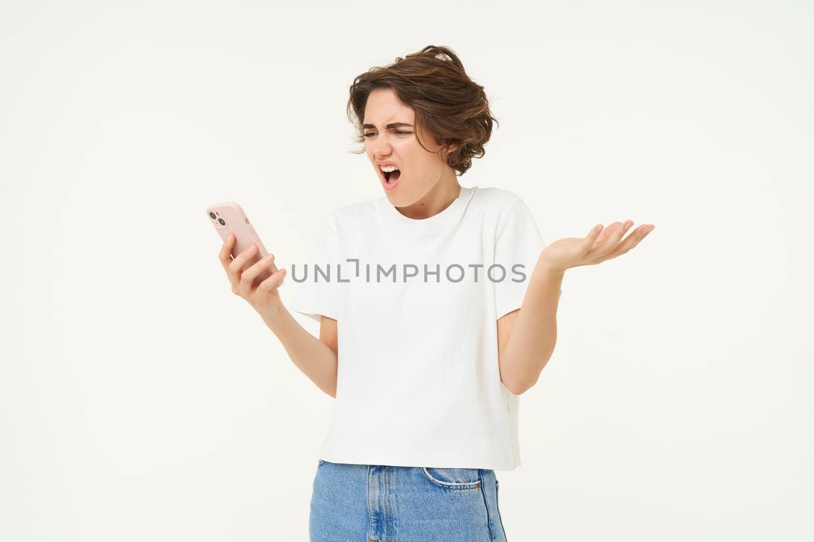 Portrait of frustrated girl with smartphone, woman looking at telephone and reacting shocked at mobile phone, shrugging shoulders, standing over white background.