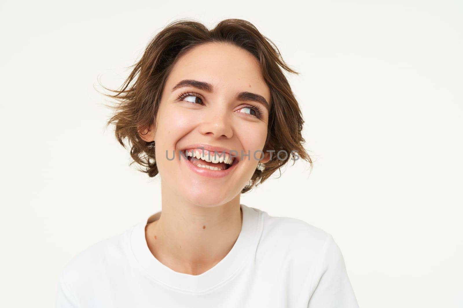 Close up shot of brunette girl with short hairstyle with genuine emotions, smiling and looking happy, standing over white background. Copy space