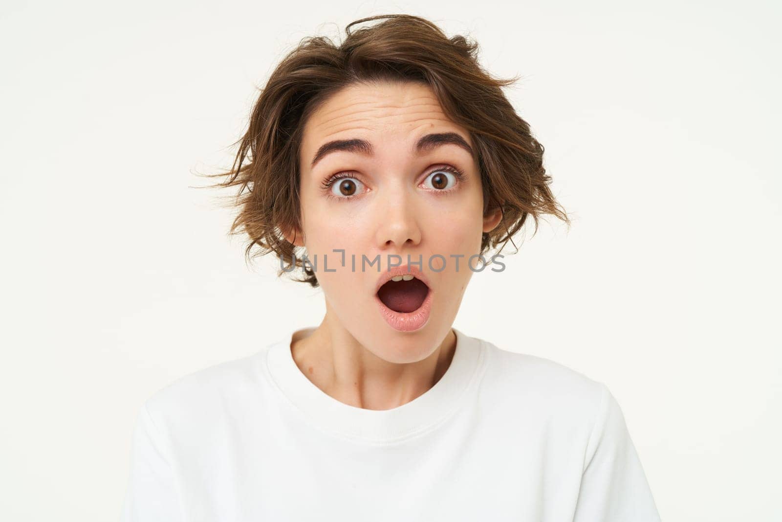 Portrait of woman in shock, drops jaw and stares at camera with mouth wide open, amazed by something, genuine surprise, stands over white background.