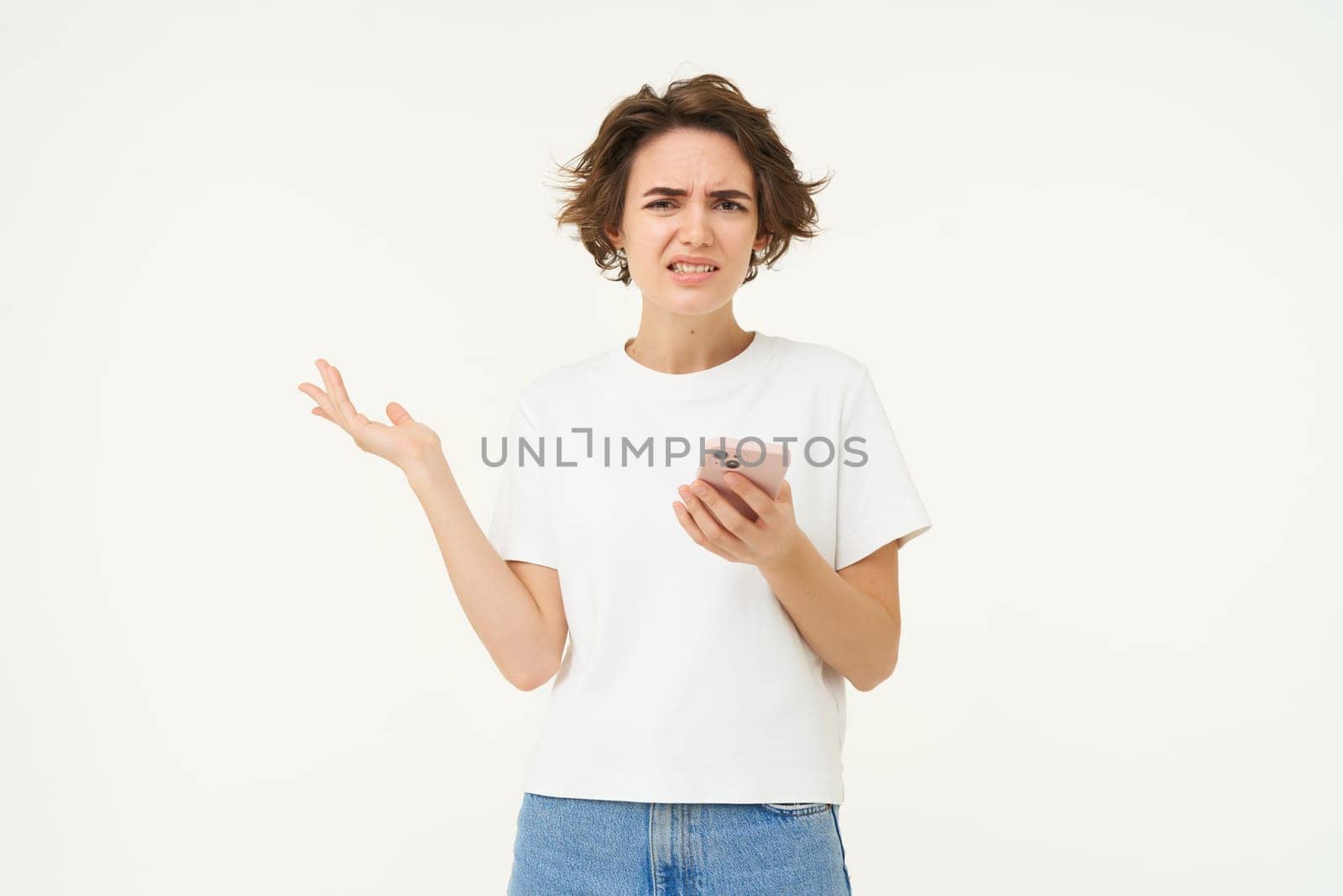 Portrait of woman with smartphone standing confused, puzzled, cant understand something online, using mobile phone, posing over white studio background.