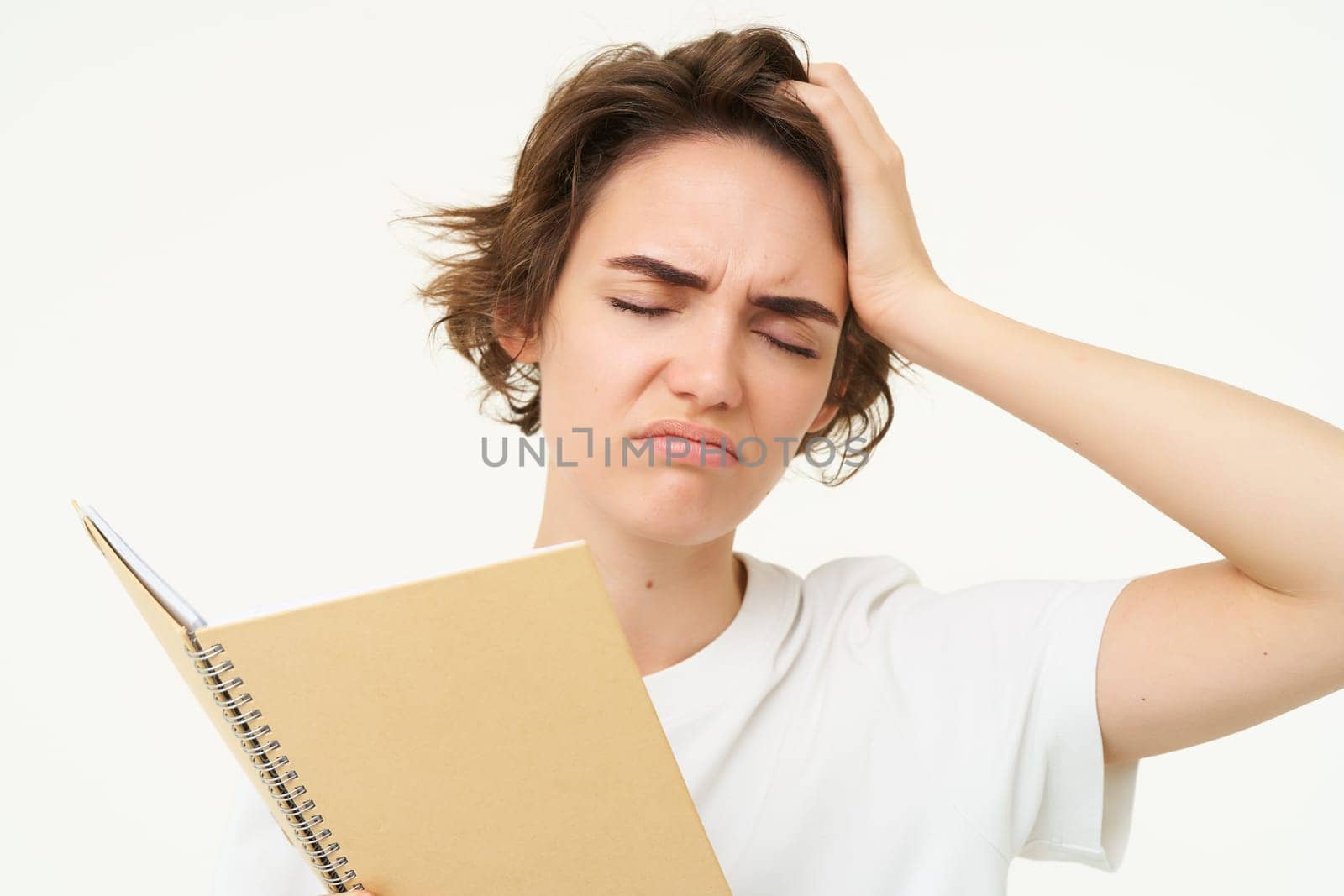 Portrait of puzzled, upset young woman tired of doing homework, holding planner with complicated face, standing over white background.