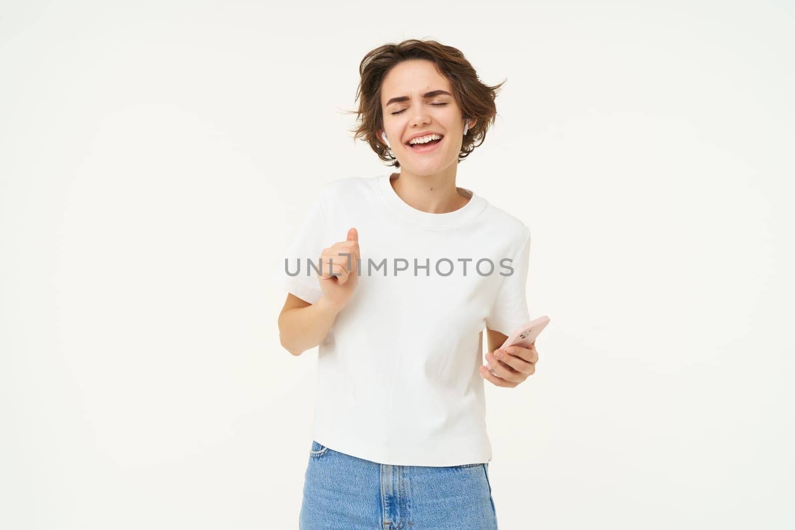 Portrait of woman singing and listening music in wireless headphones, holding mobile phone, having fun, posing over white studio background.