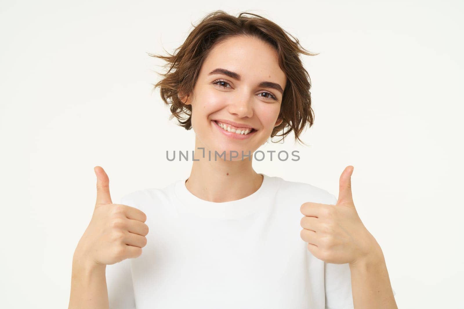 Close up of confident, smiling candid girl, shows thumbs up, recommends, gives positive feedback, approves smth good, stands over white studio background.