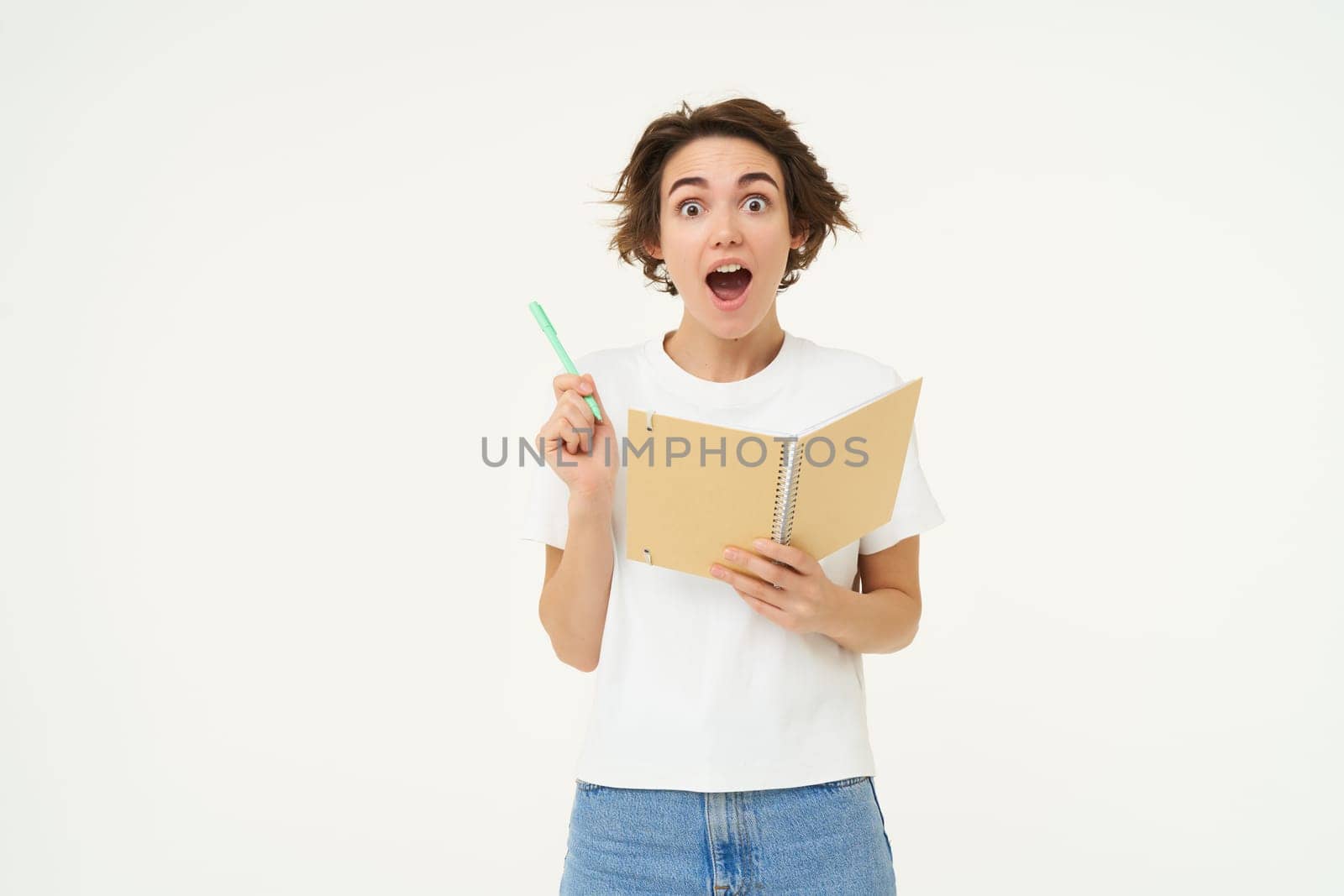 Excited young woman writing down in planner, thinking of solution, says eureka, suggests plan, stands over white studio background. Copy space