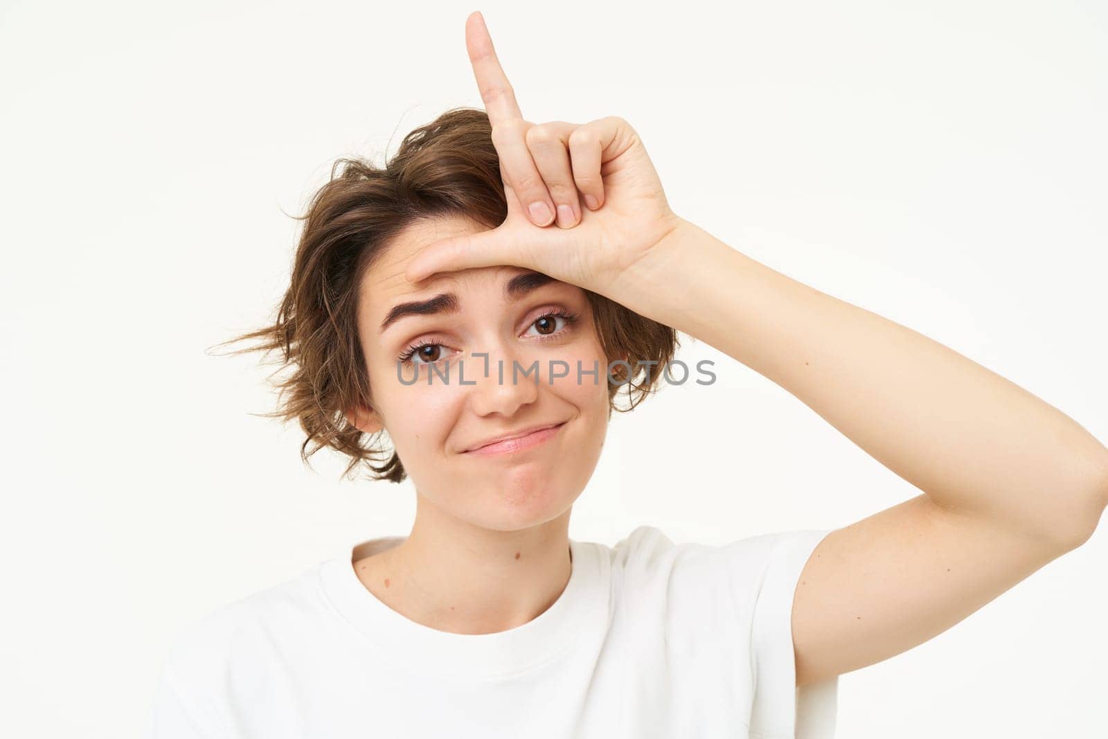 Close up of woman shows loser gesture, l letter on forehead and smiling, mocking, makes fun of someone, standing over white background.