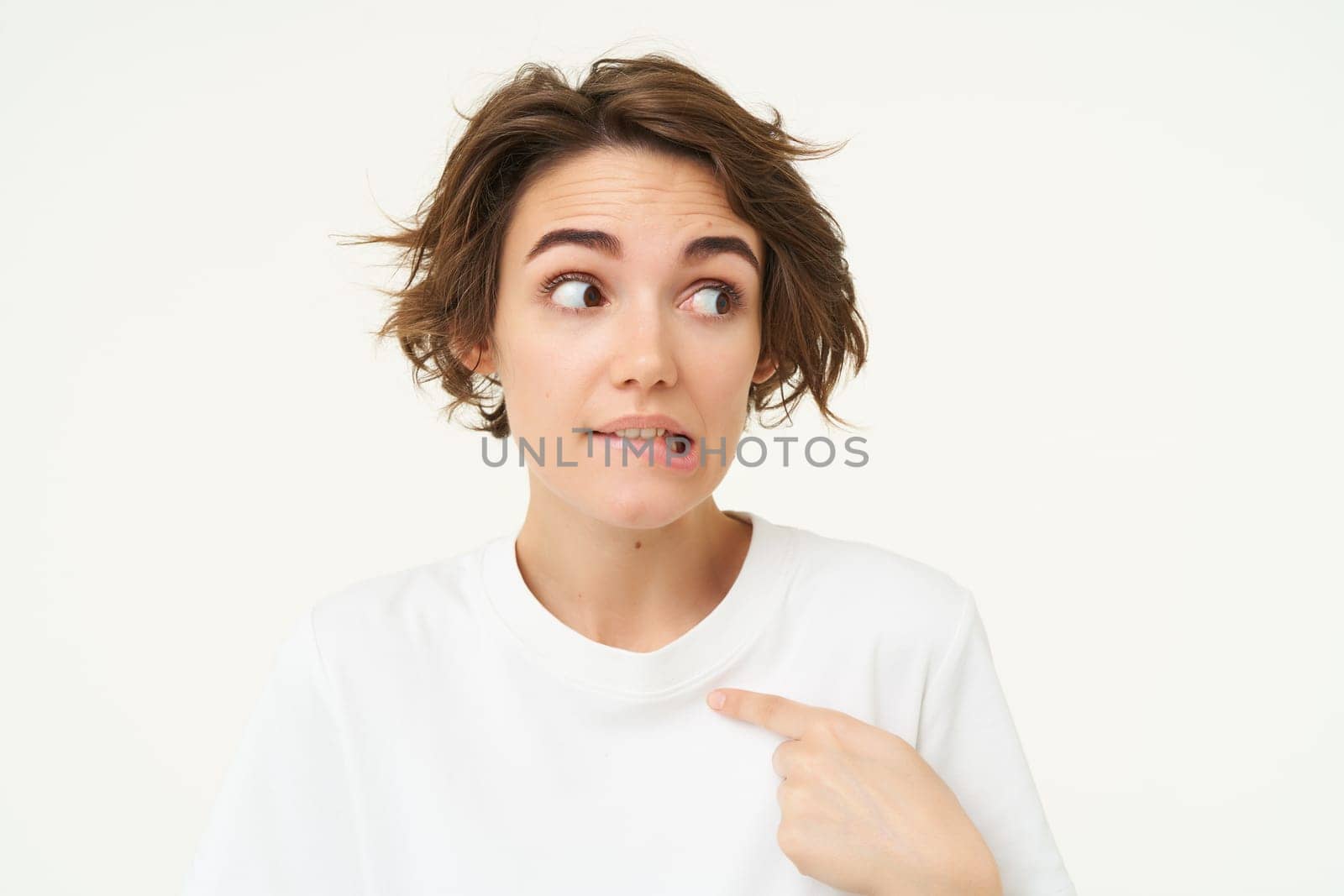 Awkward girl points at herself with guilty look, oops face, stands over white background.