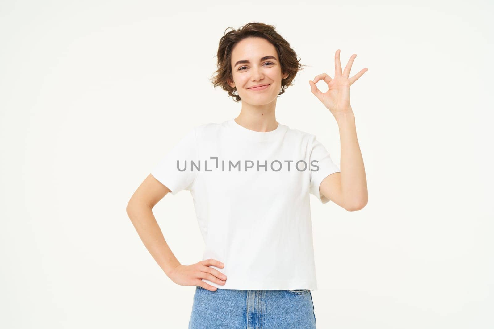 Portrait of woman smiling, showing okay sign with confidence, gives approval, recommends smth good, stands over white background.