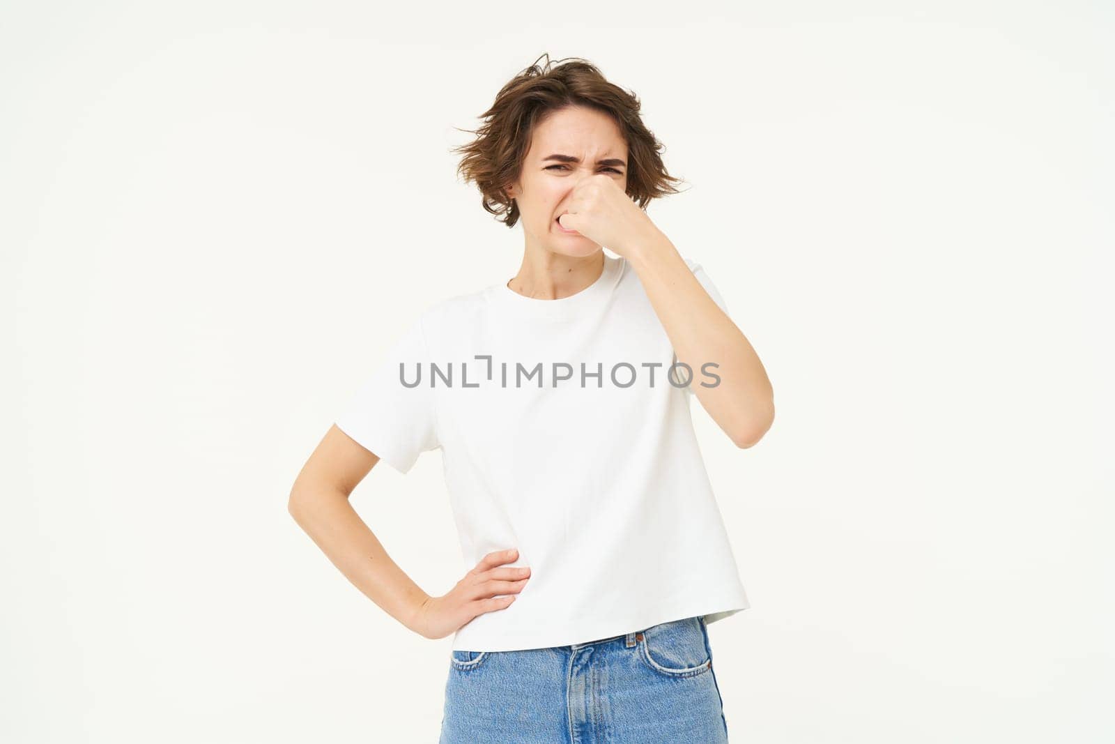 Portrait of woman disgusted by bad smell, shuts her nose, stands over white studio background.