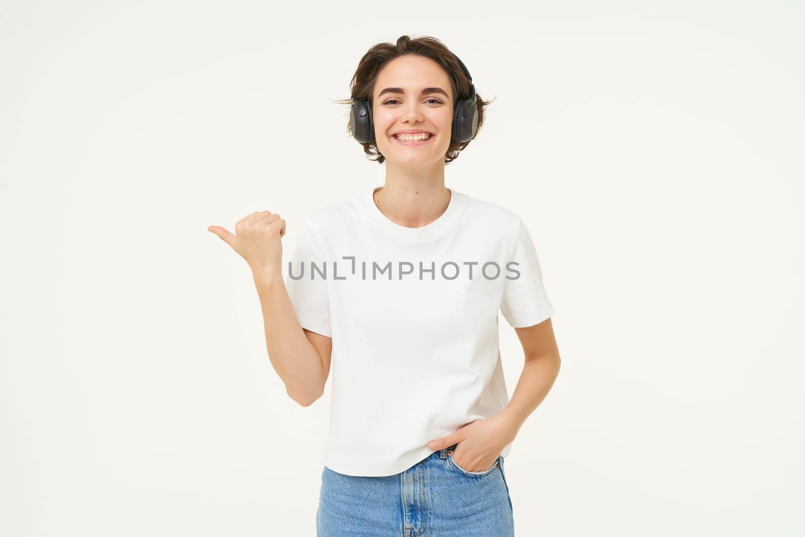 Image of young modern woman, wearing white t-shirt, pointing left, smiling and looking happy, showing advertisement, standing over white background.