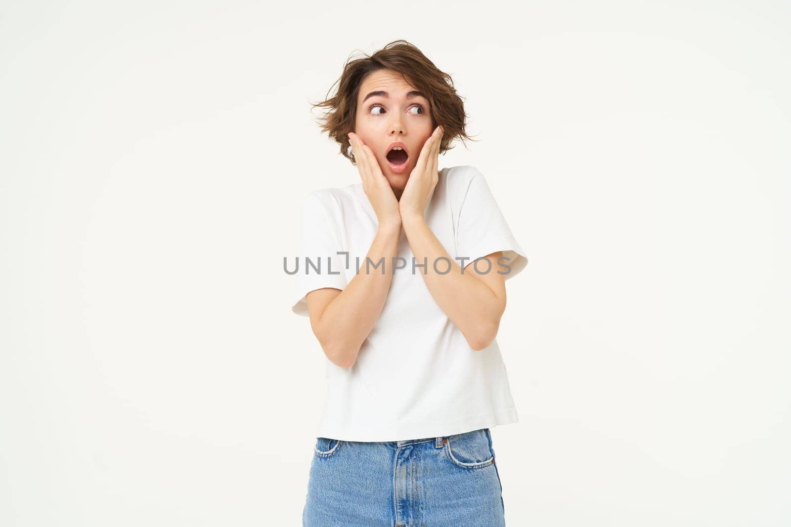 Portrait of screaming girl, holding hands on face, looks shocked and startled by something terrifying, standing over white background. copy space