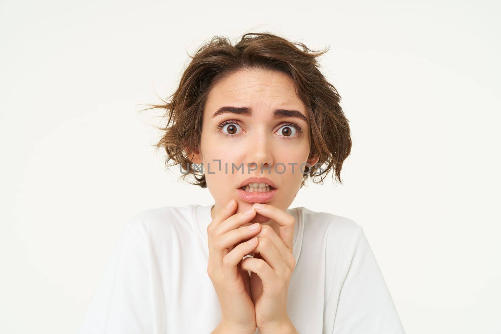 Close up of scared brunette woman, shaking from fear, looking concerned and frightened, standing over white studio background.