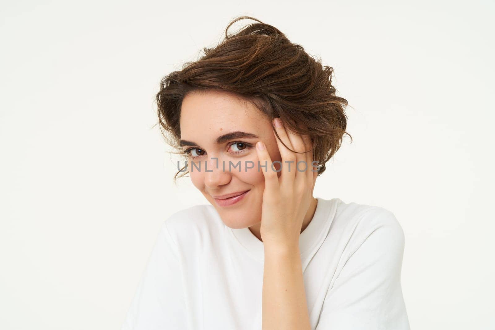 Close up of brunette woman looks shy, covers face with hand and smiles, gazing flirty at camera, stands over white background.