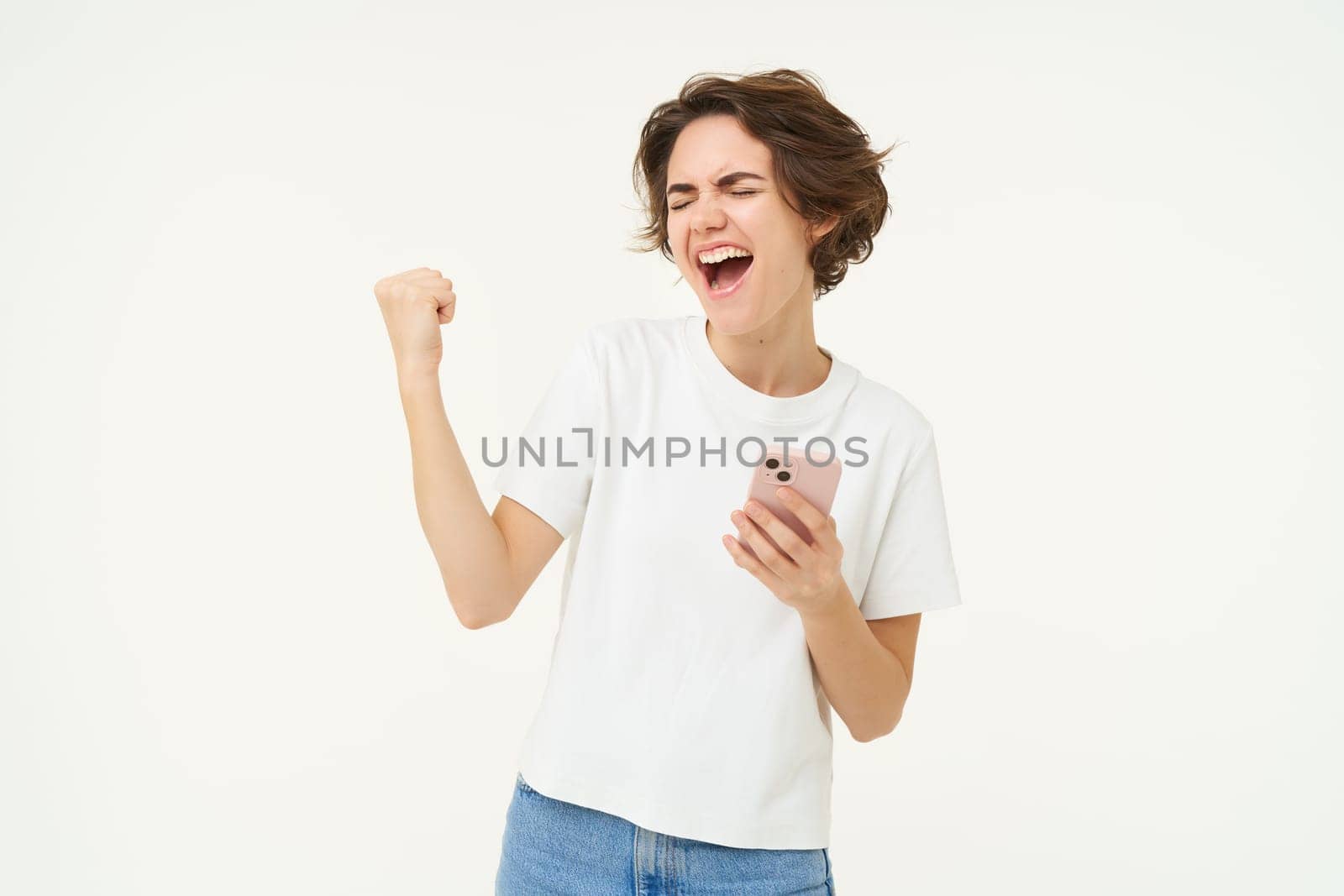 Portrait of brunette woman holding mobile phone, winning, triumphing after completing achievement on smartphone app, standing over white background.
